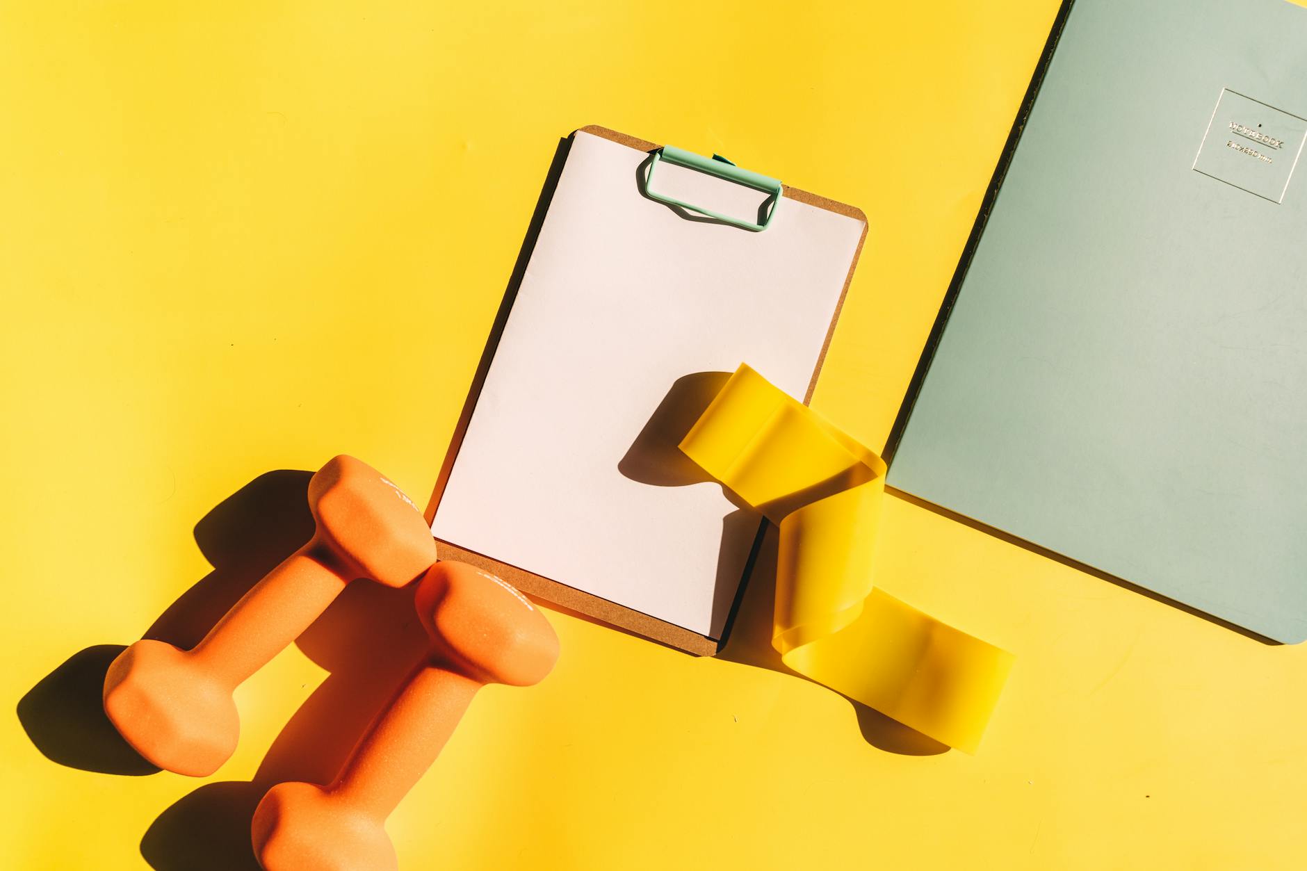 Colorful fitness setup with dumbbells, clipboard, and push straps on a vibrant background. (Photo by Alesia  Kozik on Pexels)
