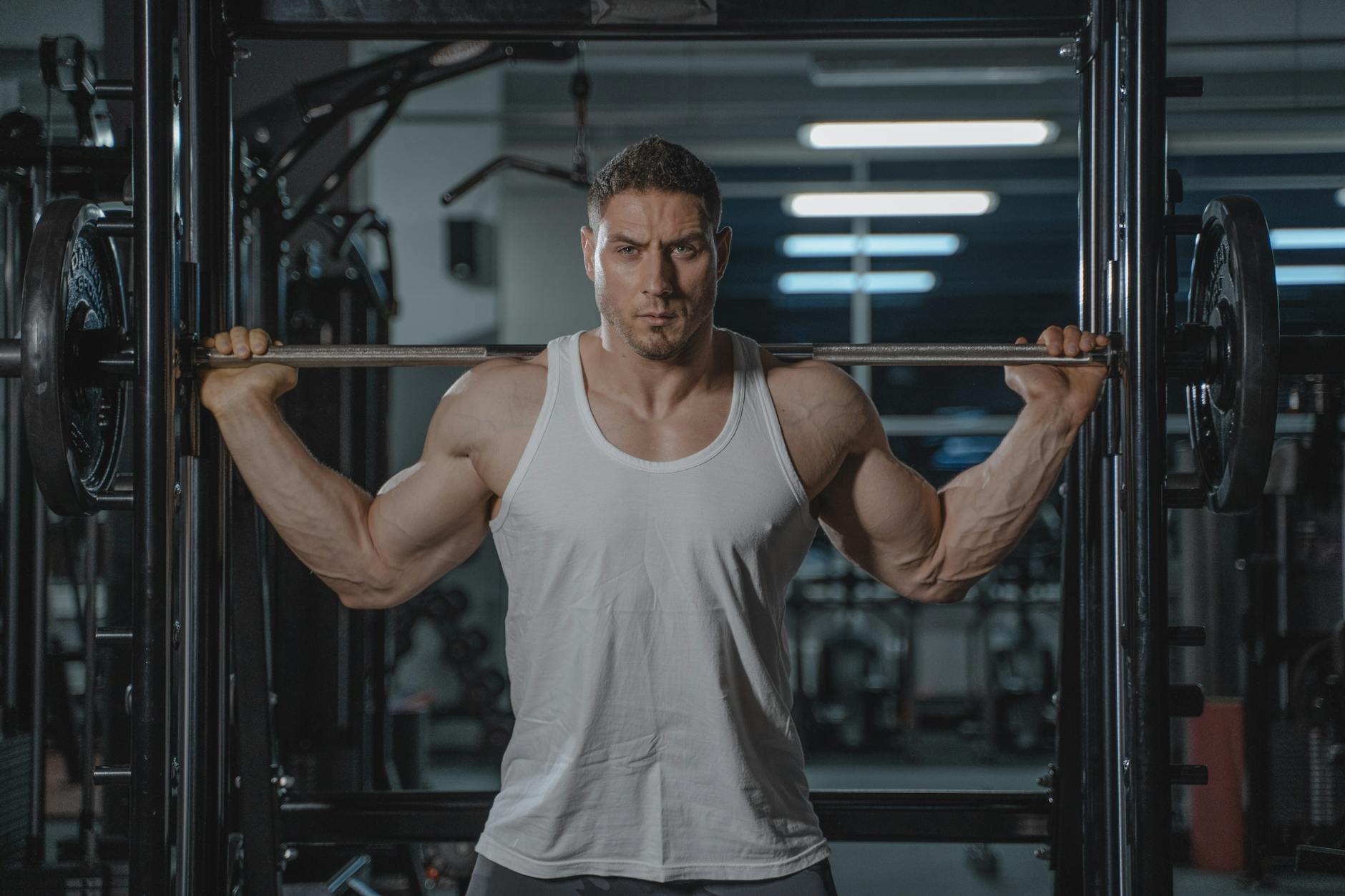 A strong, muscular man in a white tank top lifting weights in the gym. (Photo by Tima Miroshnichenko on Pexels)